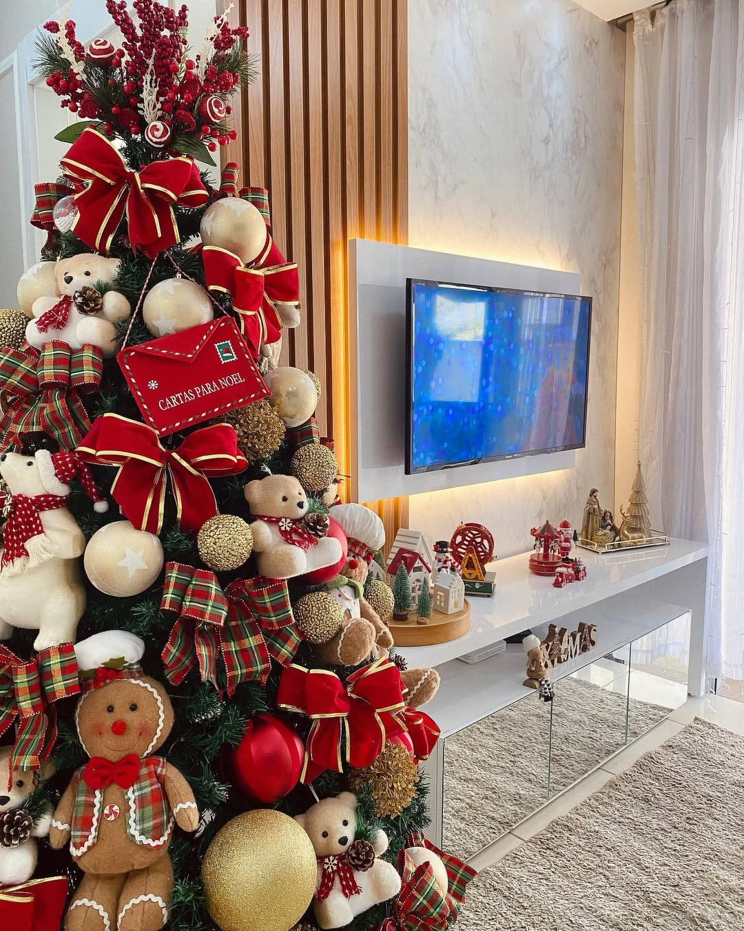 Árvore de Natal com ursinhos de pelúcia, laços vermelhos e envelope “Cartas para Noel” ao lado de uma sala decorada com mini vilarejos natalinos.