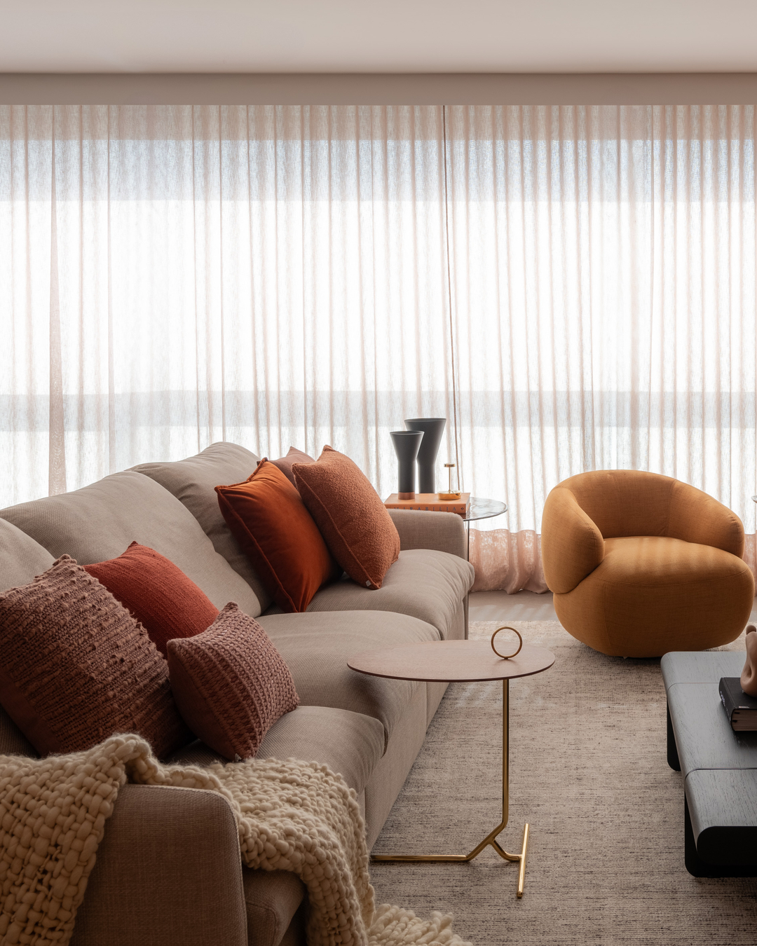 Sofá bege com almofadas terracota e poltrona mostarda em sala de estar iluminada por cortinas de linho.