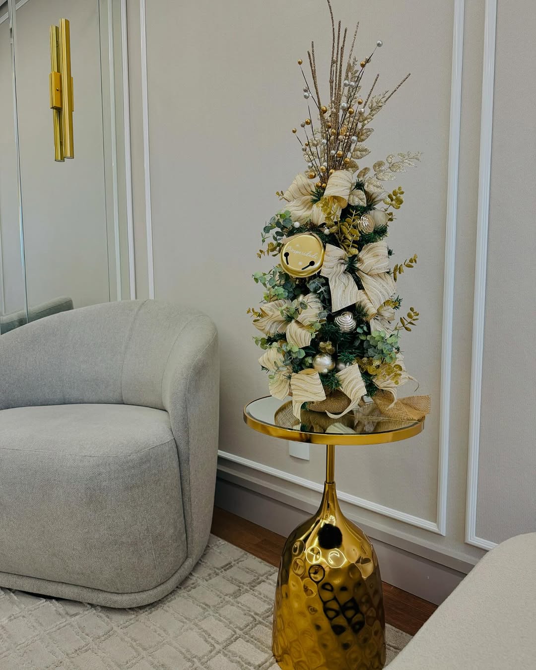 Mini árvore de Natal com laços dourados e decoração delicada sobre mesa lateral dourada espelhada em ambiente sofisticado.