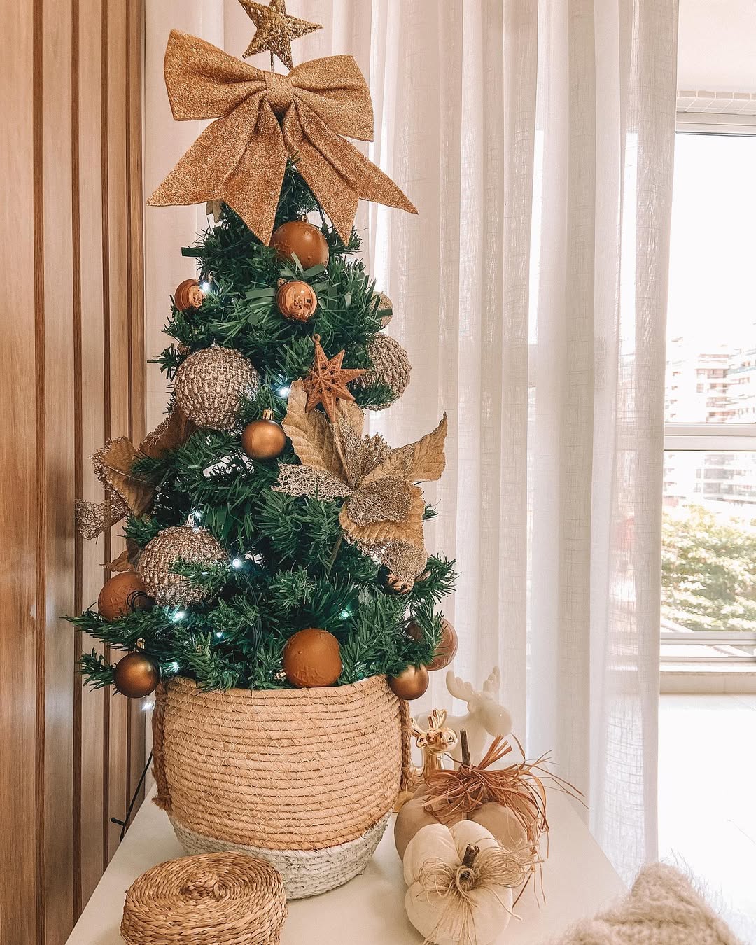 Mini árvore de Natal rústica com cesto de corda, enfeites em tons terrosos e estrelas douradas em frente à janela iluminada.