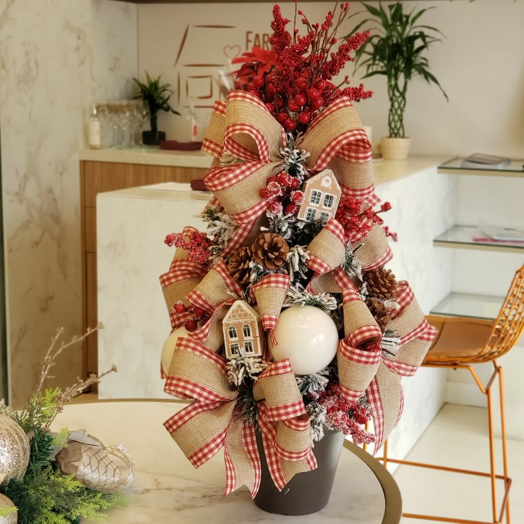 Mini árvore de Natal com laços xadrez, pinhas e casinhas decorativas em estilo rústico sobre mesa em ambiente iluminado.