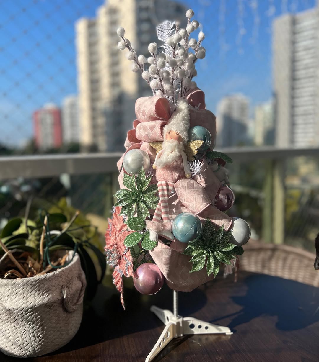 Mini árvore de Natal rosa com boneca de lã, laços acetinados e enfeites em tons pastel em varanda com fundo urbano.