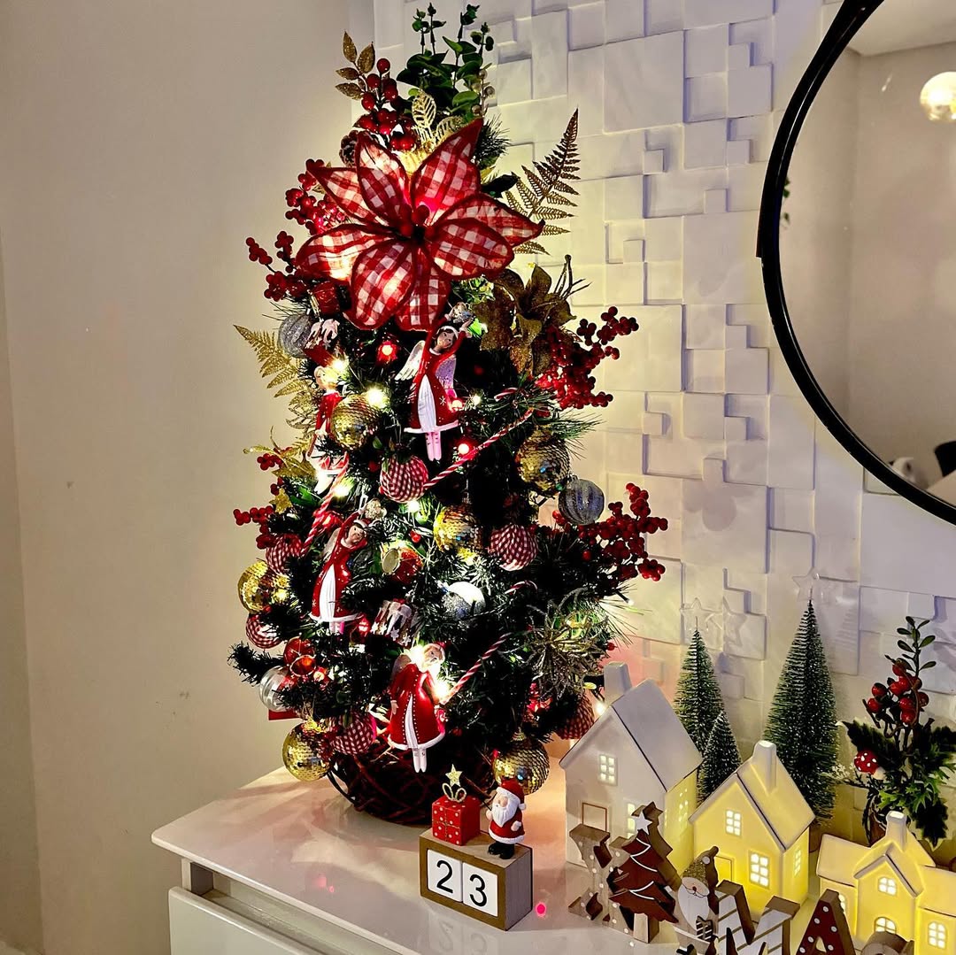 Mini árvore de Natal com laços vermelhos e dourados, ramos vermelhos e luzes sobre aparador decorado com casas iluminadas.