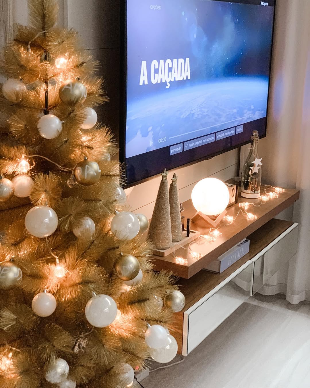 Árvore de Natal dourada com bolas brancas e douradas ao lado de rack com luzes suaves, pequenos enfeites e uma TV ao fundo exibindo o título “A Caçada”, em uma sala com decoração natalina minimalista.