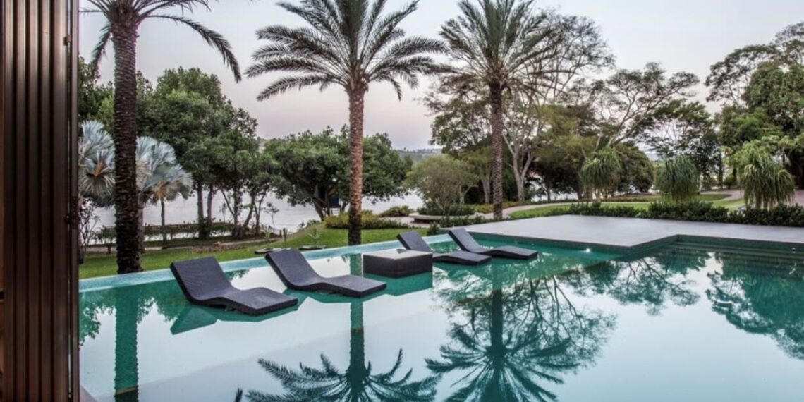 Na composição do paisagismo desta residência localizada em Brasília, os arredores da piscina revelam um amplo gramado verde e espécies como palmeiras, árvores, forrações e arbustos. Para desfrutar um clima ainda mais leve e descontraído, a prainha molhada entrega a impressão que as espreguiçadeiras estão flutuando sobre a água | Projeto de paisagismo: Depieri Paisagismo | Projeto de arquitetura: MAAI Arquitetura | Foto: Clausem Bonifacio