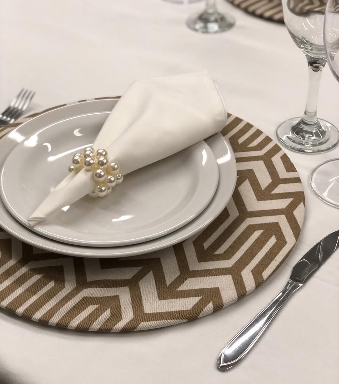 Guardanapo branco com porta-guardanapo de pérolas em prato branco sobre sousplat bege geométrico.