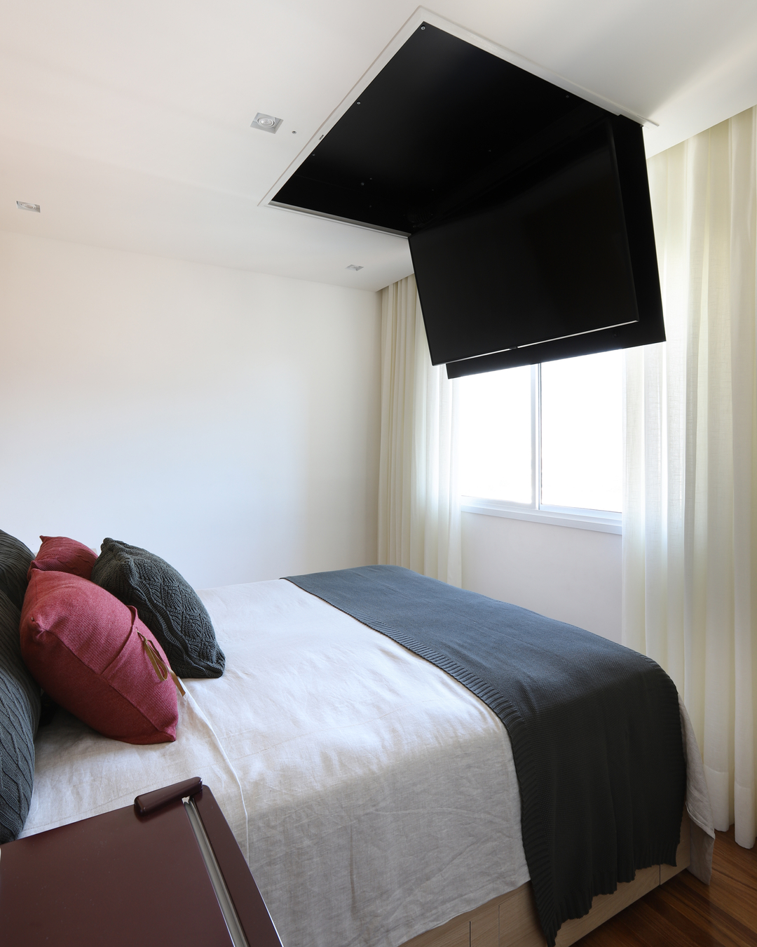 Quarto com cama de casal, cortinas claras e TV retrátil embutida no teto.