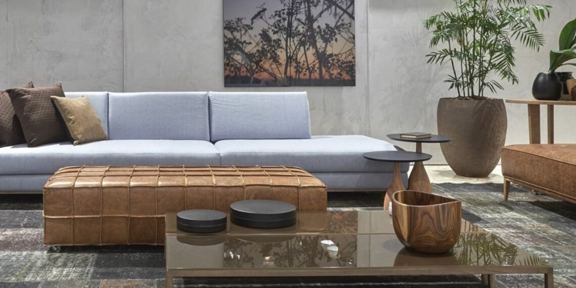 A sala revela como o estilo industrial pode ganhar sutileza sem perder personalidade. O cimento queimado das paredes cria o pano de fundo perfeito para o sofá de linhas retas em tom azul — uma escolha que suaviza a estética urbana sem diluir sua força visual. O pufe em couro natural envelhecido e a mesa de centro com estrutura metálica reforçam a combinação entre materiais brutos e conforto, enquanto o quadro com paisagem ao entardecer introduz um toque poético ao ambiente. A grande planta no vaso rústico equilibra o concreto com frescor, reforçando o conceito contemporâneo de industrial acolhedor.