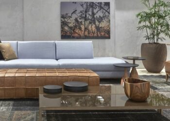 A sala revela como o estilo industrial pode ganhar sutileza sem perder personalidade. O cimento queimado das paredes cria o pano de fundo perfeito para o sofá de linhas retas em tom azul — uma escolha que suaviza a estética urbana sem diluir sua força visual. O pufe em couro natural envelhecido e a mesa de centro com estrutura metálica reforçam a combinação entre materiais brutos e conforto, enquanto o quadro com paisagem ao entardecer introduz um toque poético ao ambiente. A grande planta no vaso rústico equilibra o concreto com frescor, reforçando o conceito contemporâneo de industrial acolhedor.