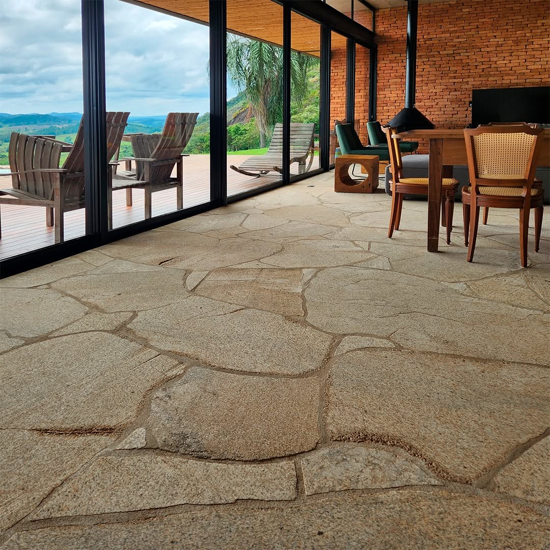 Interior de sala com piso de pedra moledo em tons beges, compondo com móveis de madeira e parede de tijolos, ao lado de ampla esquadria de vidro com vista para área externa com deck.