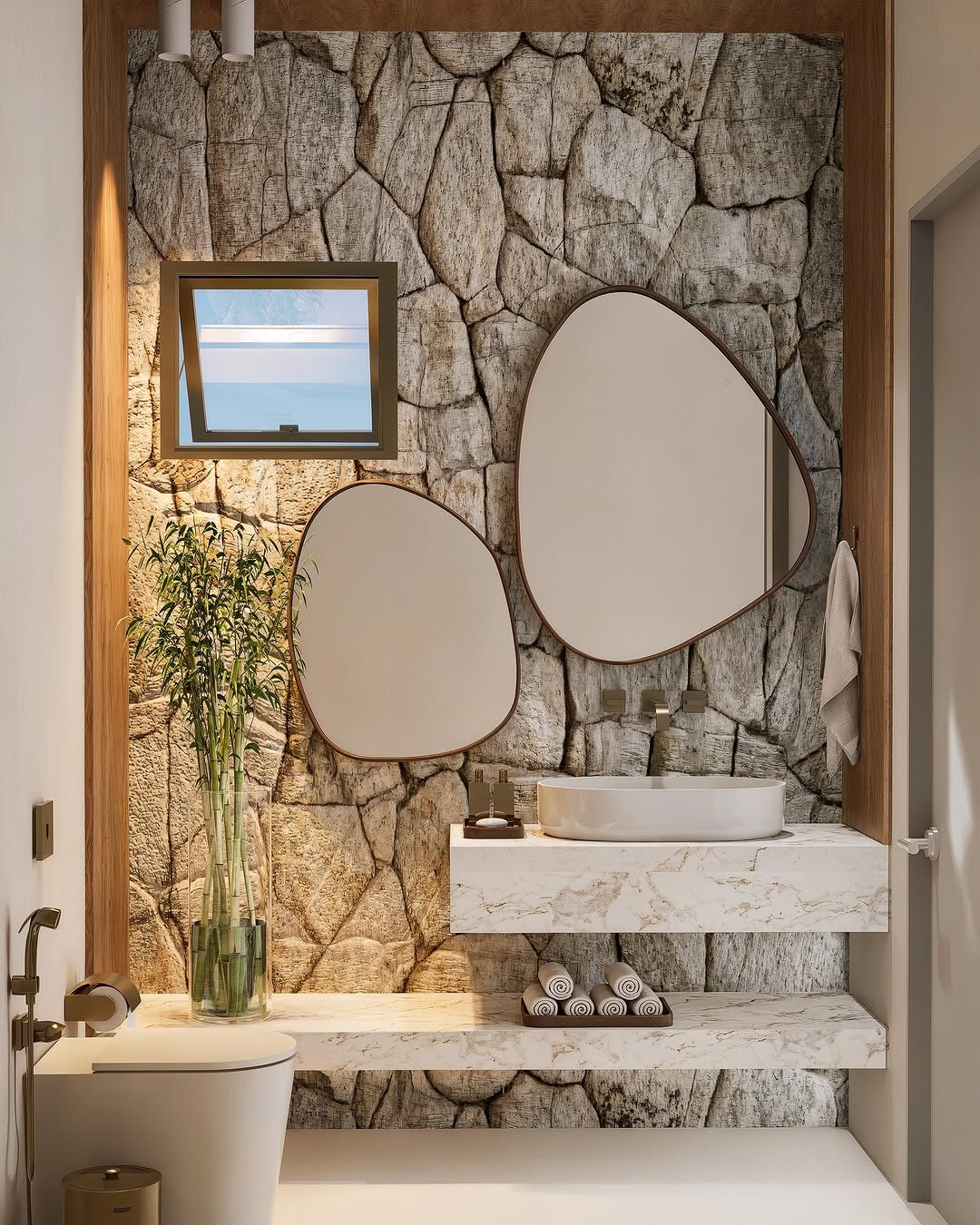 Lavabo moderno com parede revestida de pedra moledo, espelhos orgânicos, bancada de mármore e detalhes em madeira, criando um ambiente elegante e acolhedor.