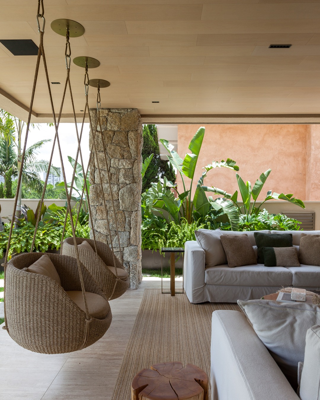 Varanda coberta com sofá claro e três poltronas-balanço de fibra penduradas no teto, ao lado de pilar de pedra e jardim de plantas tropicais, formando uma área de estar aconchegante.