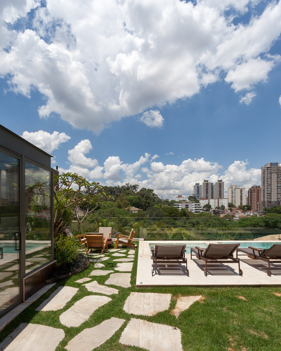 Jardim com caminho de pedras que leva a um deck com espreguiçadeiras ao lado da piscina, cercado por vegetação e com vista para prédios e área verde ao fundo.