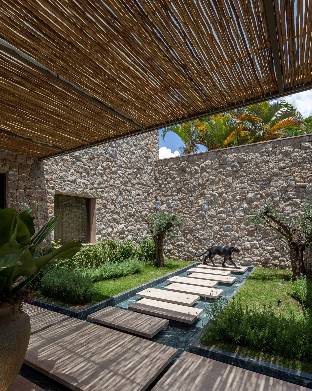 Pátio com piso de placas sobre espelho d’água, jardim gramado com arbustos, paredes de pedra e escultura de pantera, sob pergolado de ripas de madeira que projeta sombras no chão.