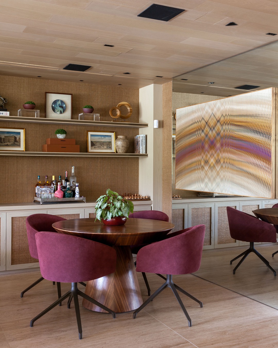 Sala com mesa redonda de madeira, quatro cadeiras de veludo vinho, prateleiras com objetos decorativos, bar discreto e grande obra de arte colorida ao fundo, em ambiente revestido com fibras naturais.