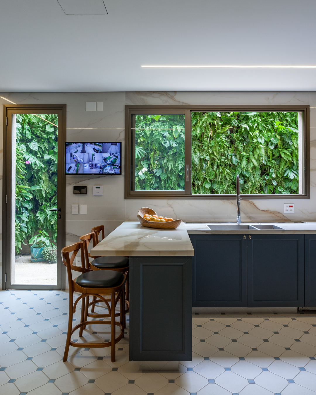 Cozinha com ilha e armários azul petróleo, bancadas em pedra clara, piso geométrico e grandes janelas voltadas para um jardim vertical verde, com banquetas de madeira encostadas na ilha.