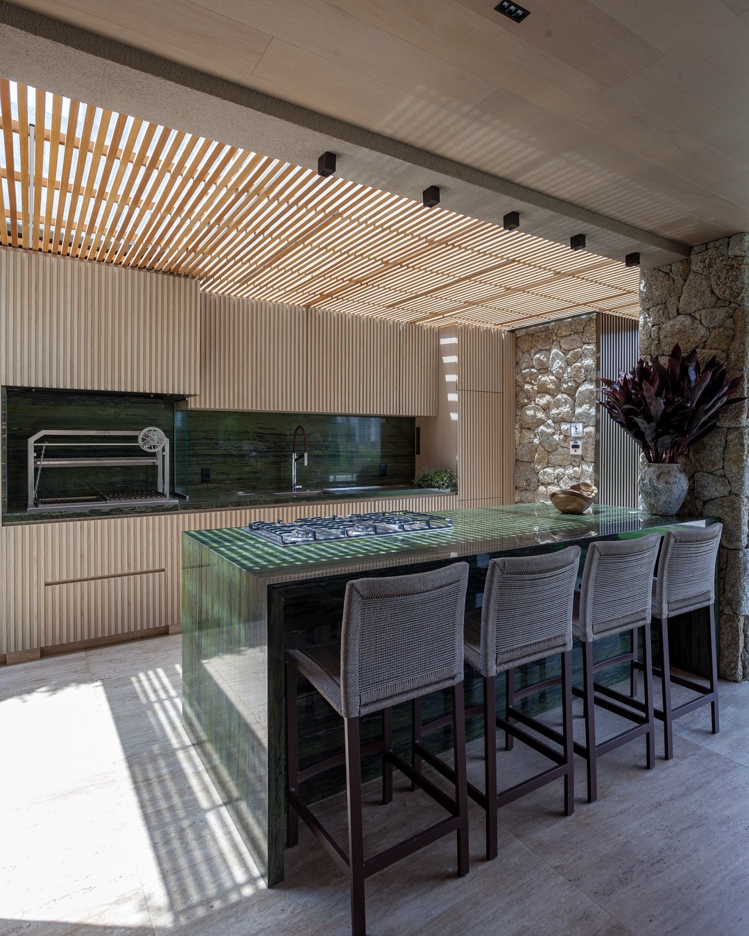 Área gourmet com ilha em pedra verde, cooktop embutido, quatro banquetas altas, marcenaria ripada clara, parede de pedras e cobertura ripada que projeta sombras no piso iluminado.