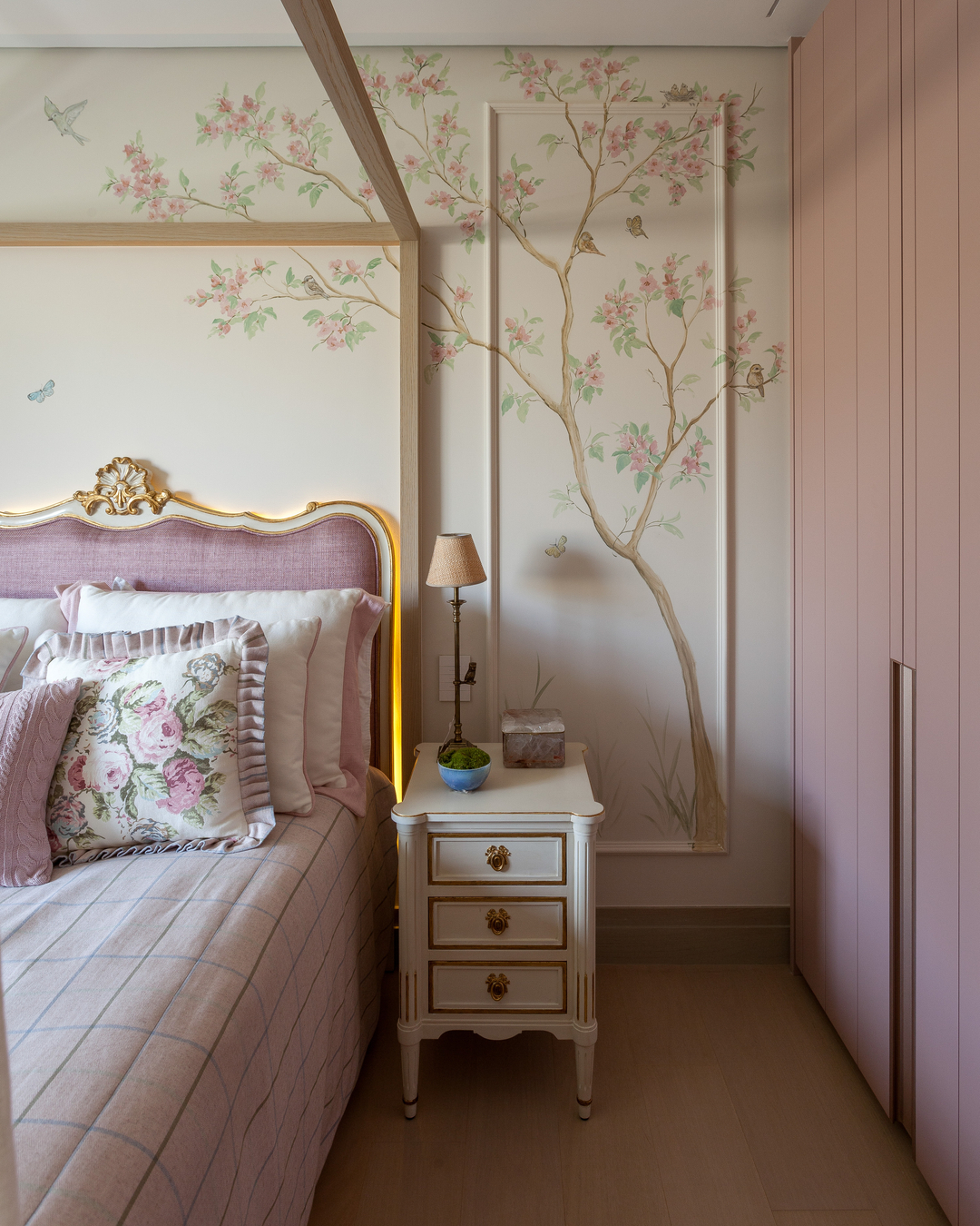 Quarto feminino com cama de dossel rosé, criado-mudo clássico branco e parede com mural floral pintado à mão em tons suaves de rosa e verde, ao lado de armário planejado rosa-claro.