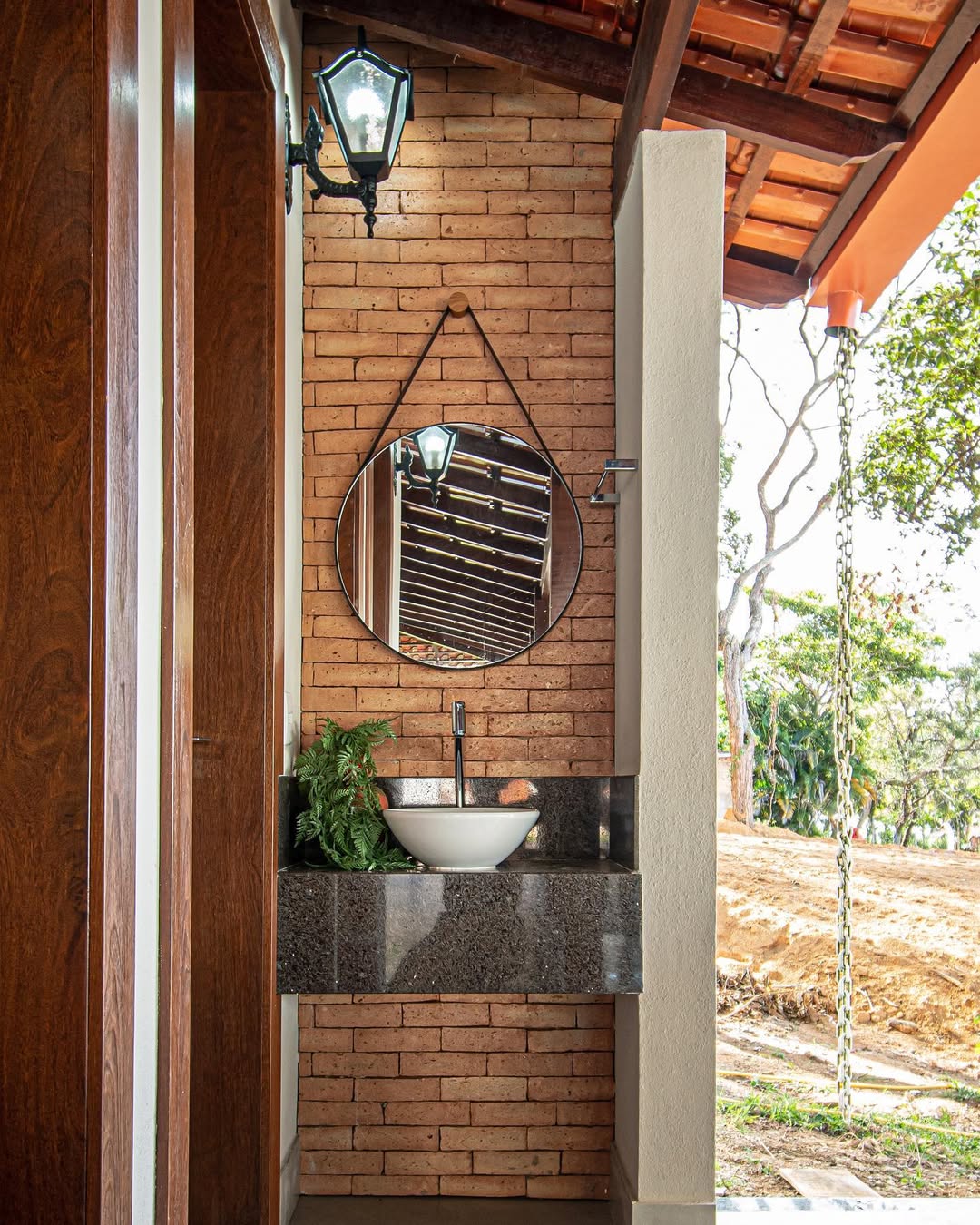 Lavabo externo com parede de tijolinhos, bancada em granito escuro, cuba branca redonda e espelho redondo pendurado com fita, decorado com planta verde ao lado da pia.