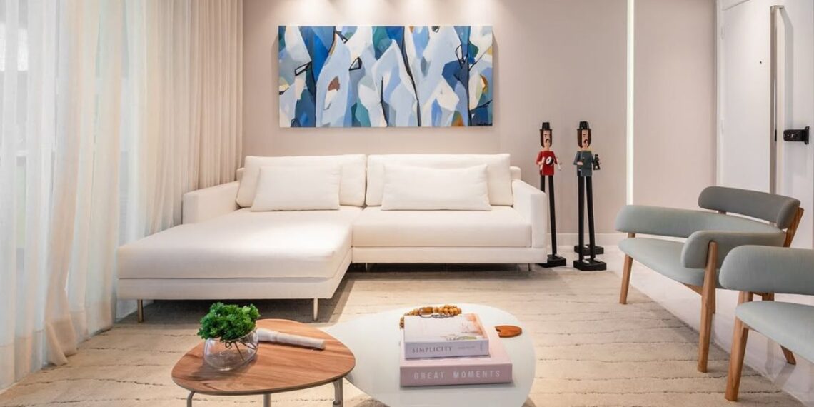 A sala ganha um ar de leveza com o sofá branco em formato lounge, que dialoga com a paleta suave das paredes e com a obra abstrata em tons de azul. As mesas de centro, em formas orgânicas, criam um ponto de encontro acolhedor, enquanto os bonecos escultóricos dão personalidade e charme ao ambiente. | Imagem: gabipontualarquitetura