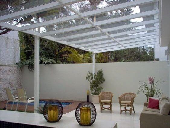 Pergolado de vidro com estrutura branca em área externa, poltronas em fibras naturais, decoração minimalista e integração com jardim, criando ambiente sofisticado, iluminado e aconchegante.
