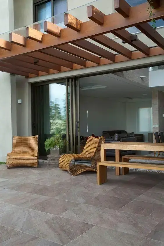 Pergolado de vidro com estrutura em madeira sobre área externa integrada à sala, espreguiçadeiras de fibra natural, mesa de madeira e piso em pedra, criando ambiente sofisticado, iluminado e aconchegante.