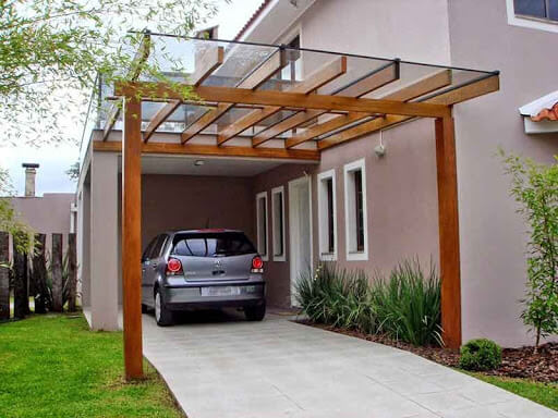 Pergolado de vidro com estrutura em madeira natural cobrindo garagem residencial, permitindo entrada de luz natural, integrado ao paisagismo e à fachada, com design moderno, funcional e acolhedor.
