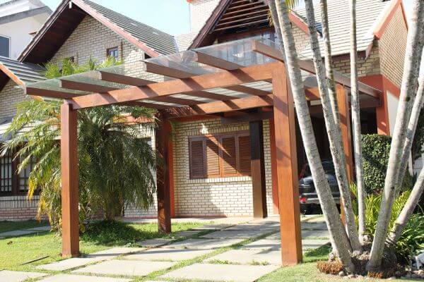 Pergolado de vidro com estrutura em madeira na entrada de casa, integrado ao paisagismo tropical e à fachada residencial, criando acesso protegido, iluminado e visualmente acolhedor.