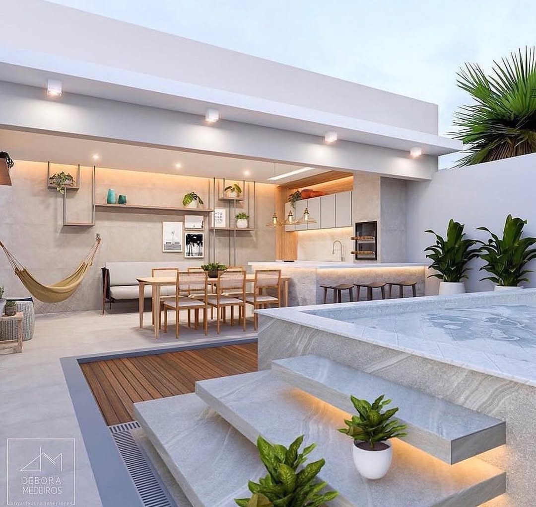 Piscina com churrasqueira integrada em área externa moderna, com deck de madeira, iluminação embutida, bancada gourmet, mesas de jantar e paisagismo, destacando integração, conforto e design contemporâneo.