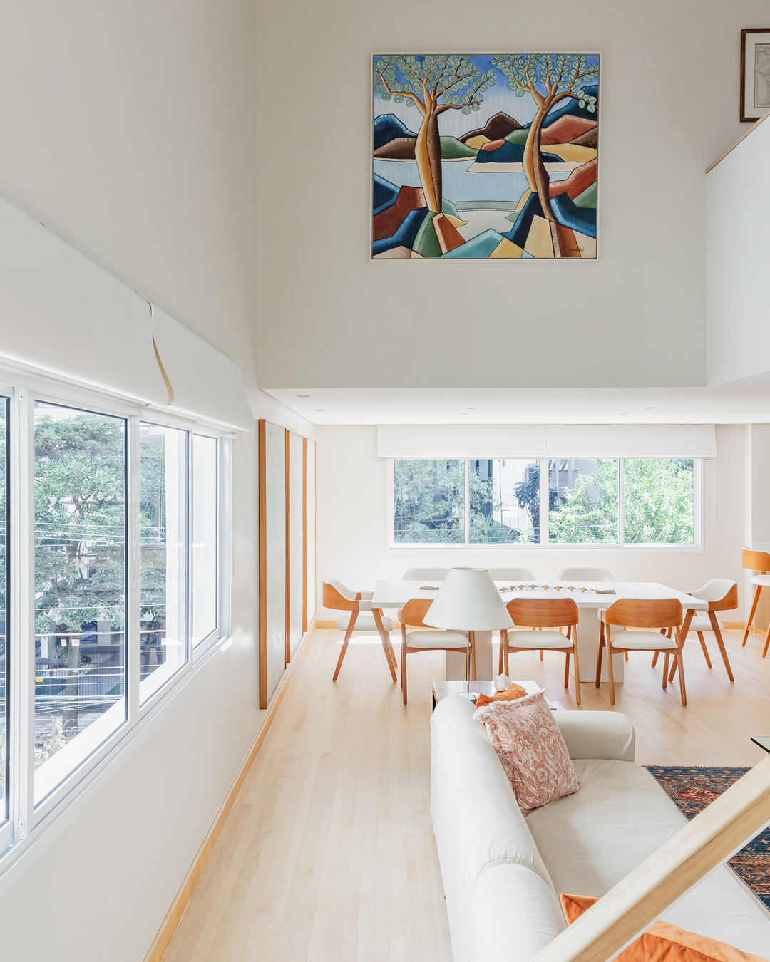 Sala de jantar com grandes janelas, mesa ampla, cadeiras em madeira clara e arte na parede, em ambiente iluminado e integrado.
