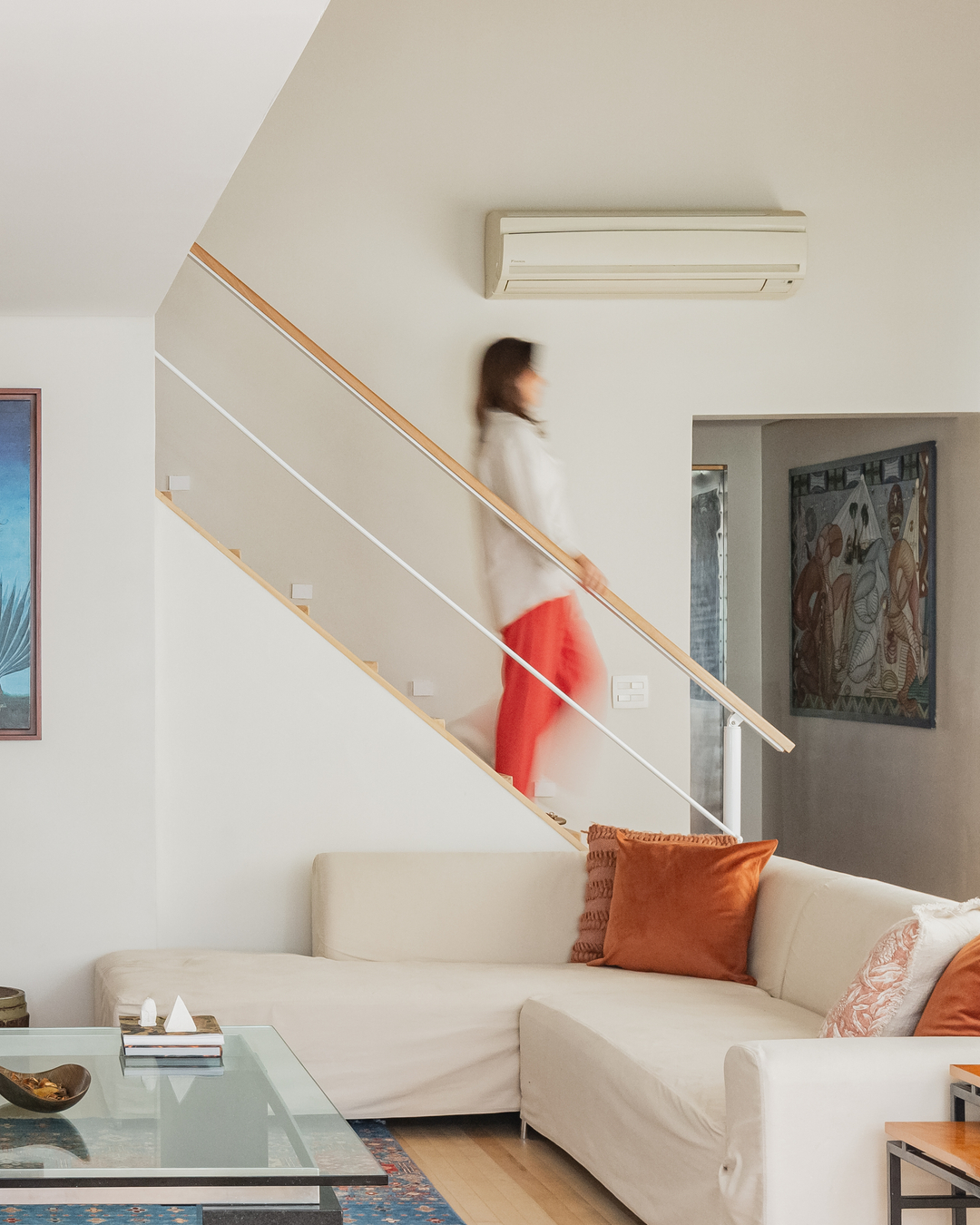 Sala de estar com sofá claro em L, almofadas terracota, escada com guarda-corpo branco e atmosfera leve, ideal para relaxar.