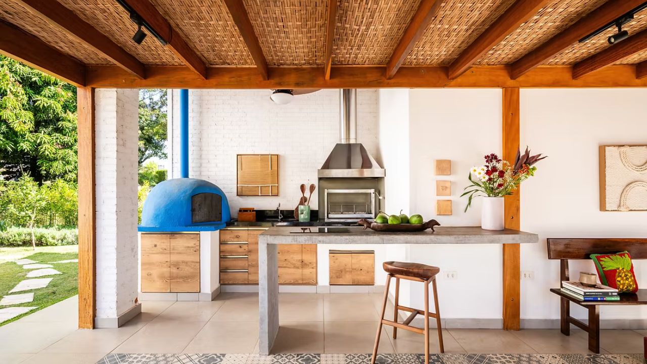 Área gourmet com bancada em concreto, forno de pizza azul, madeira natural e forro de palha em casa revitalizada.