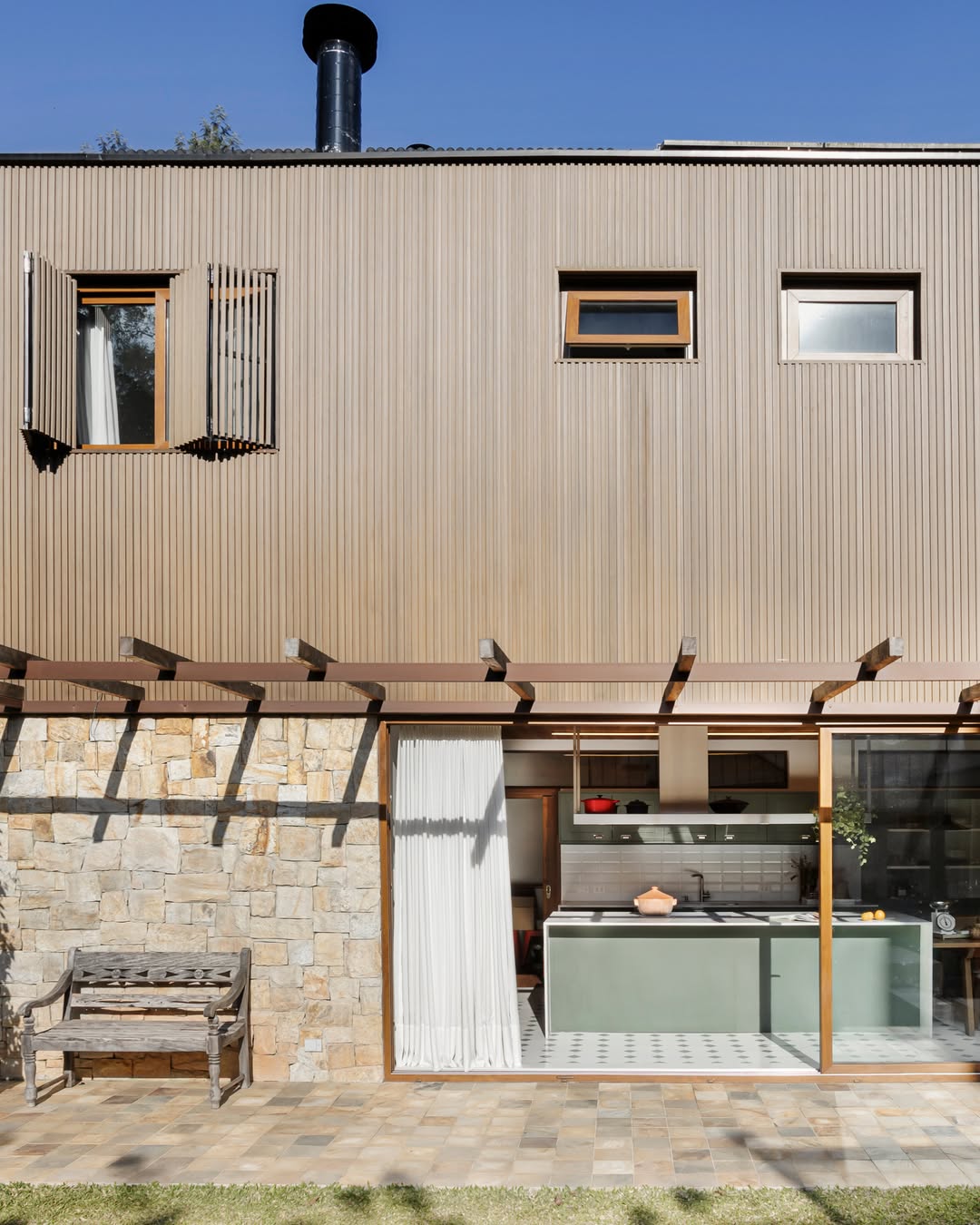 Área externa de casa em Curitiba com pérgola de madeira, piso de pedra natural e cozinha integrada ao jardim por esquadrias amplas — Estudio Elmor.