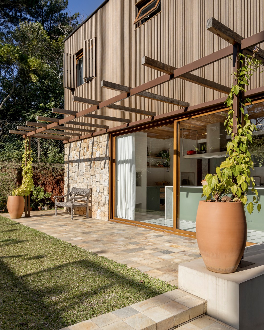 Jardim com pérgola de madeira e metal, vasos de cerâmica terracota e acesso à cozinha por esquadria de correr — residência do Estudio Elmor em Curitiba.