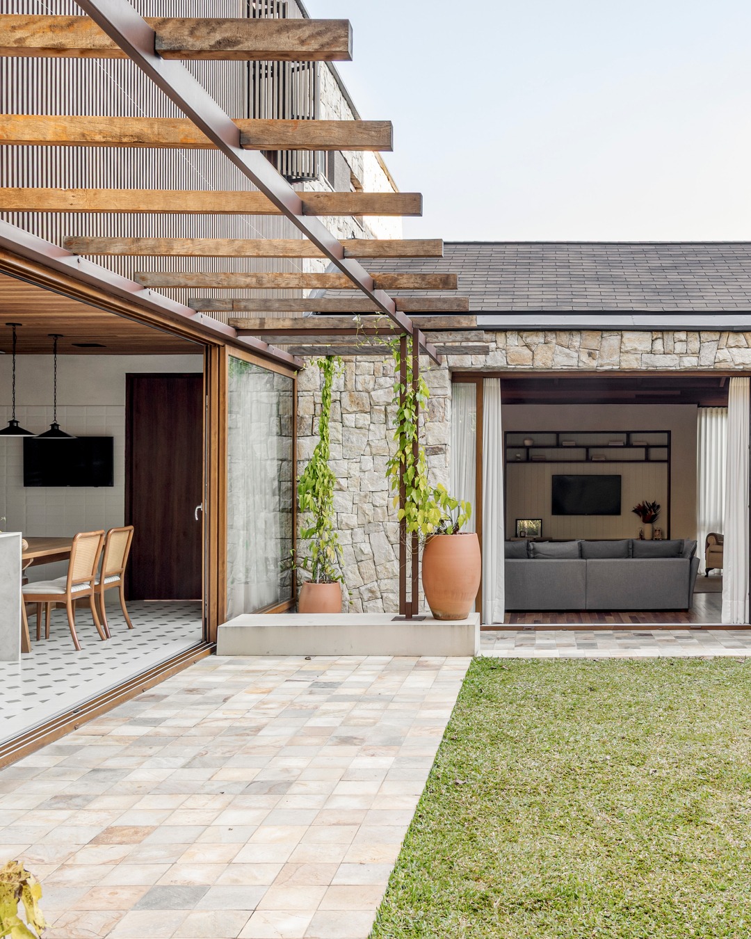 Vista do jardim para sala de estar e cozinha de casa em Curitiba, com pedra natural, pérgola de madeira e esquadrias de correr — Estudio Elmor.