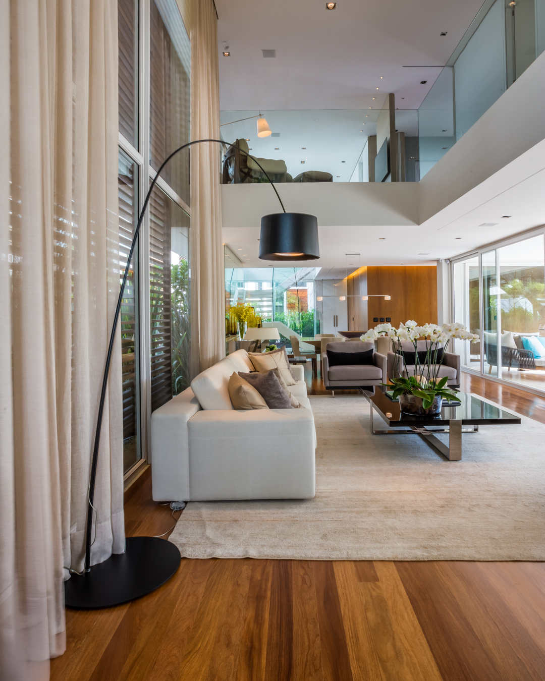 Living de casa no Brooklin Velho com piso de madeira, sofá branco, luminária de arco preta e pé-direito duplo com mezanino envidraçado ao fundo