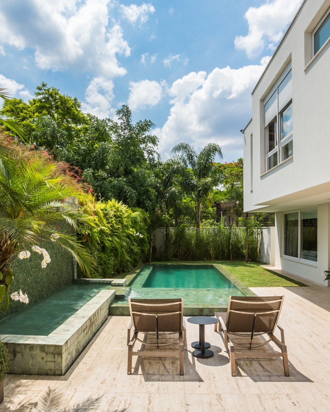 Área externa com piscina retangular revestida em pedra verde, deck em travertino, espreguiçadeiras e jardim tropical exuberante com palmeiras e samambaias