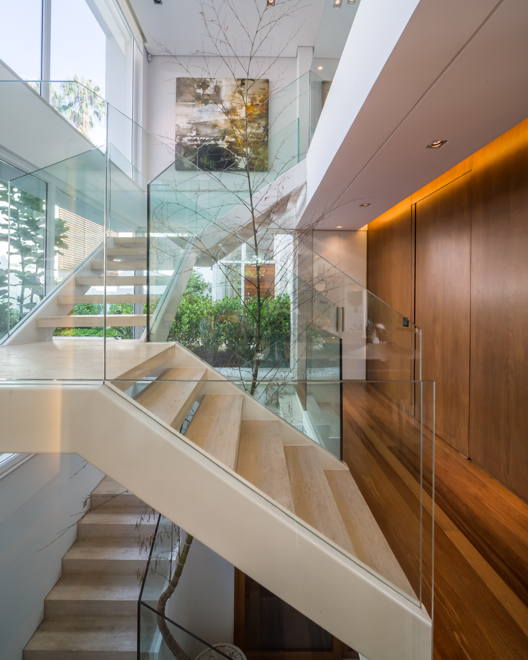 Escada escultórica de travertino com guarda-corpo de vidro, galho seco como elemento decorativo vertical e painéis de madeira na parede lateral em residência no Brooklin Velho