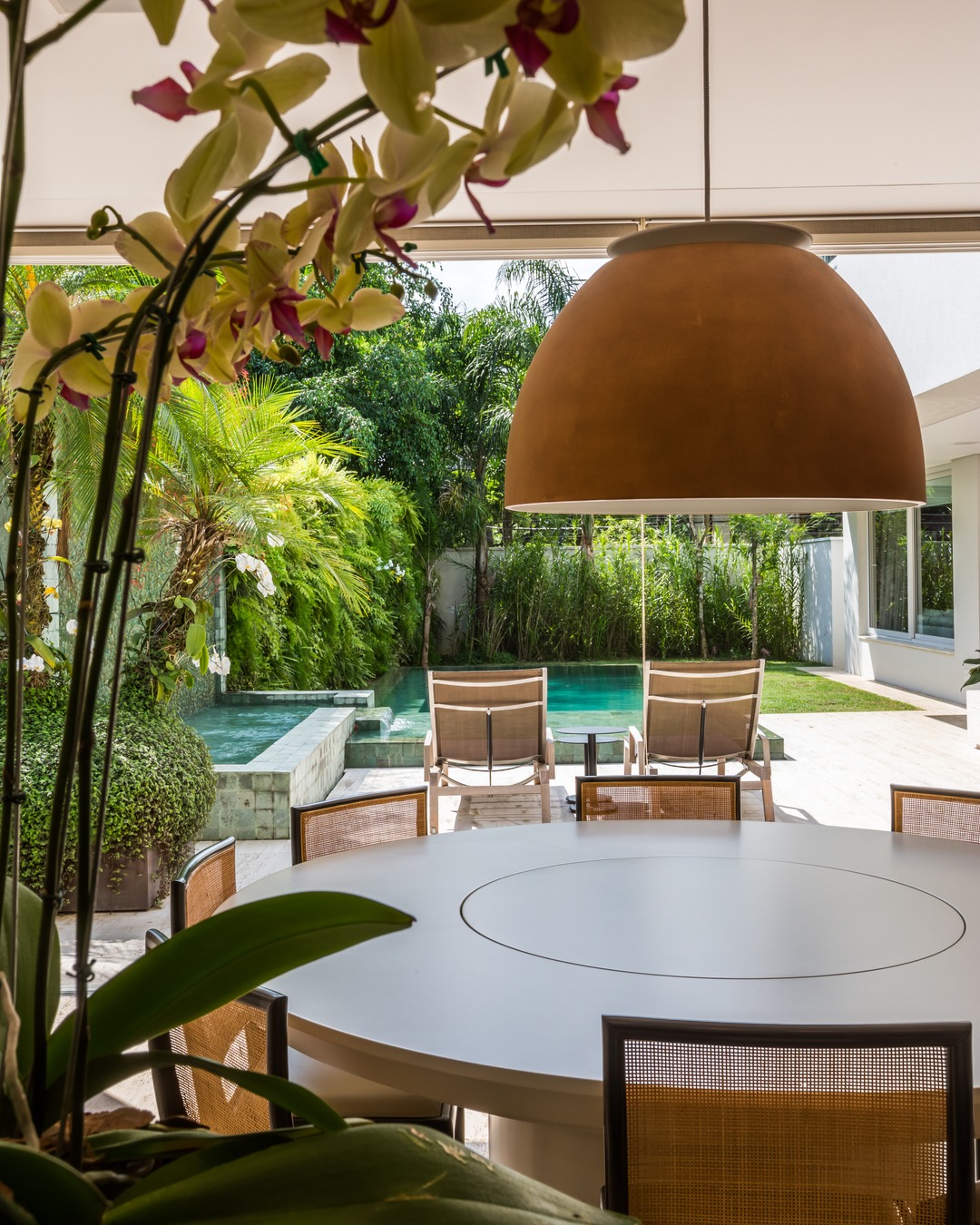 Área gourmet integrada ao jardim com mesa redonda branca, cadeiras de palha, pendente terracota e vista para piscina com paisagismo tropical em casa no Brooklin Velho, São Paulo