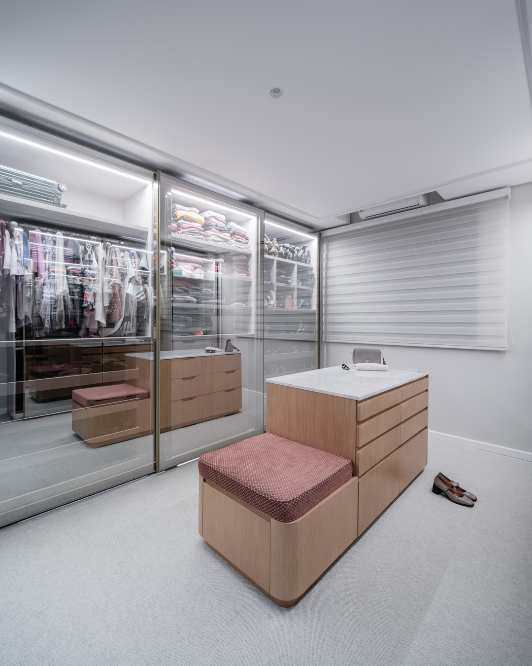 Closet com portas de vidro, iluminação interna e ilha central em madeira clara.