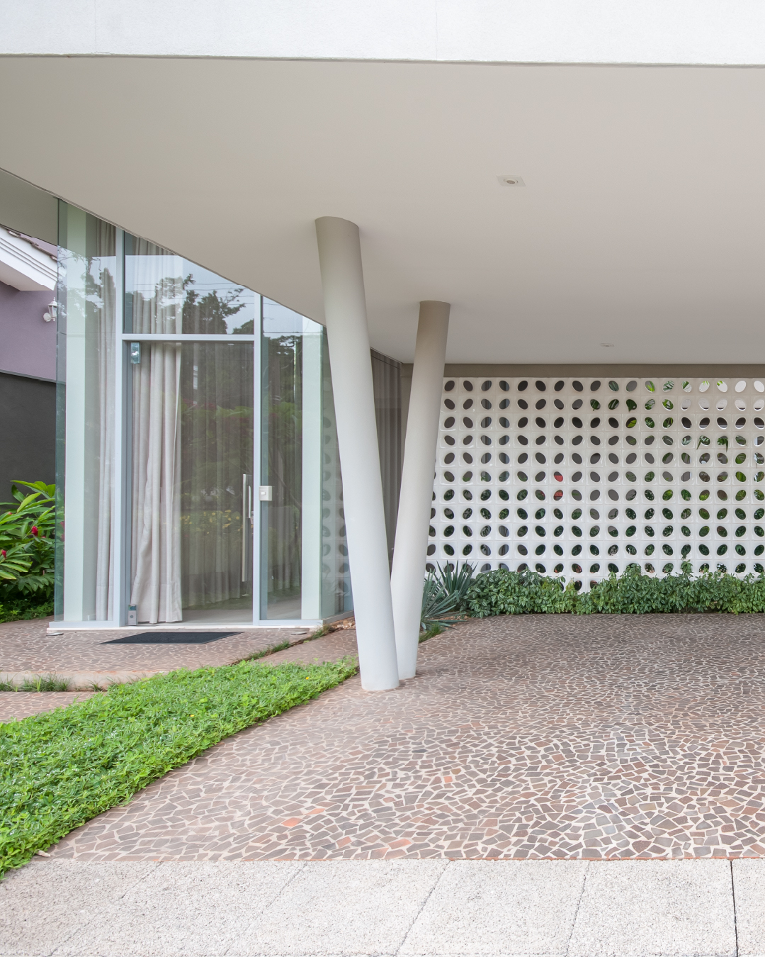 Parede de cobogó em concreto sob varanda com pilares inclinados e piso em mosaico, exemplo de arquitetura brasileira contemporânea.