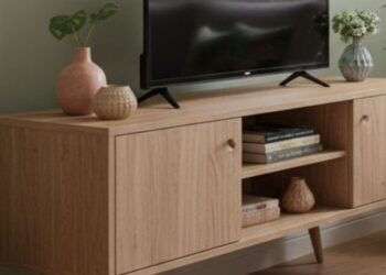 Rack pé palito em madeira clara com TV sobre o tampo, sofá bege, almofadas estampadas e plantas decorativas em sala iluminada.