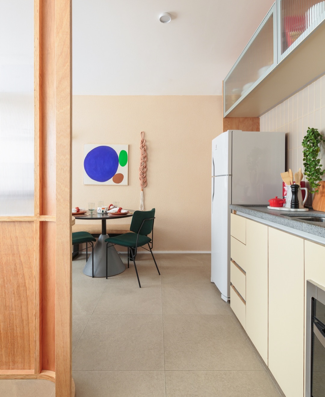 Vista do corredor entre cozinha e sala com painel de compensado naval, policarbonato leitoso e sala ao fundo com parede pêssego e quadro abstrato colorido.