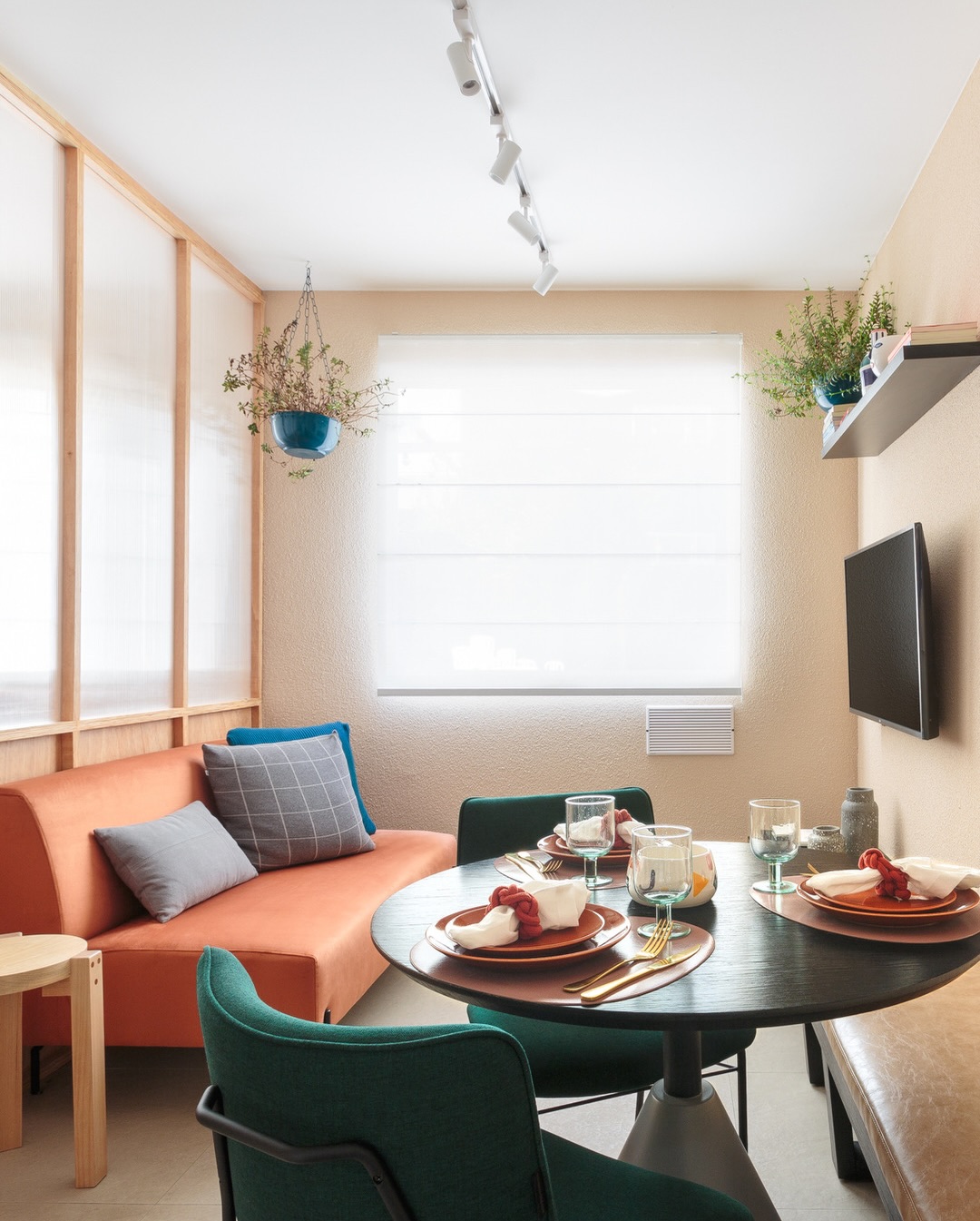 Sala integrada com sofá terracota, mesa redonda escura, cadeiras verde-musgo, vasos de plantas suspensos e trilho de spots no teto em apartamento compacto em São Paulo.
