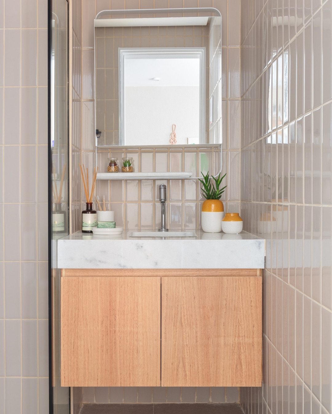 Banheiro compacto com azulejo vertical greige do piso ao teto, gabinete de madeira clara, tampo de mármore branco e espelho com moldura arredondada.