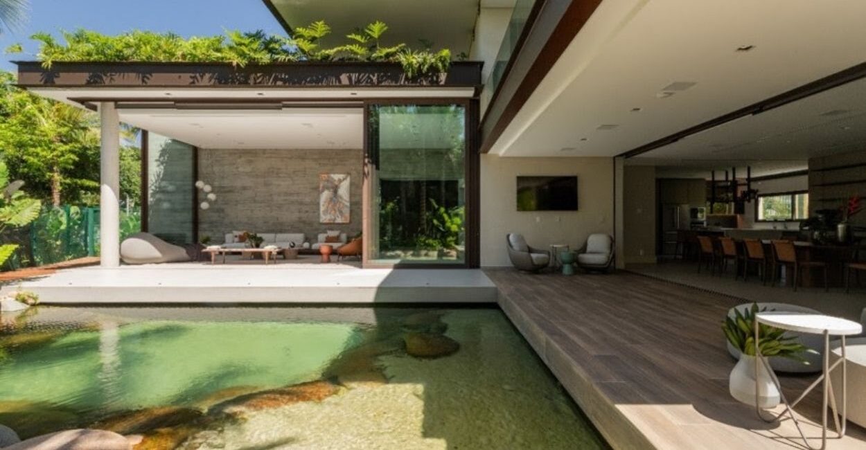 No projeto desta residência, com a assinatura da arquiteta Patricia Penna, a piscina lago se integra ao paisagismo de forma orgânica com suas bordas, pedras naturais e madeira. Atrás dele, o grande painel de vidro, com 7,20 m, se abre por completo, diluindo os limites entre o interior e o exterior| FOTO: Leandro Moraes
